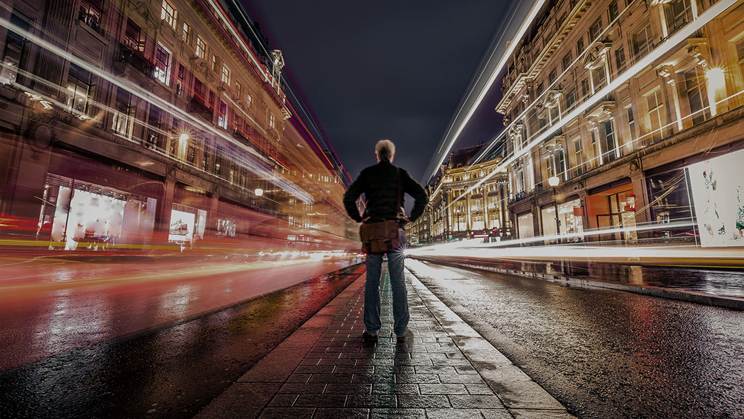 Man standing in the middle of the street at night with long exposure lights moving past him